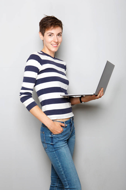 Porträt einer lächelnden jungen Frau mit Laptop vor grauem Hintergrund  - Foto, Bild
