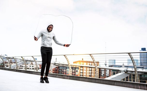Young active black sportsman skipping with a rope in a city, wearing hoodie. - Фото, изображение