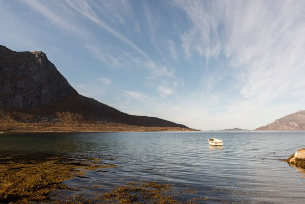 Kis, fából készült csónak kikötve a fjord, Tromso, Norvégia közelében a sziklák az előtérben és a távoli hegyek, egy alacsony, szög, kilátás a hullámok a víz a kék ég alatt - Fotó, kép