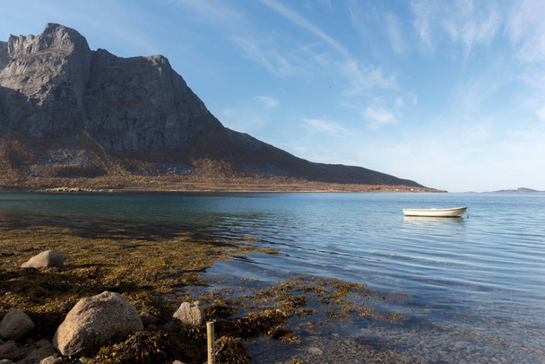 Small wooden rowboat moored in a fjord near Tromso, Norway with rocks in the foreground and distant mountains under a blue sky in a low angle view with ripples on the water - Photo, Image