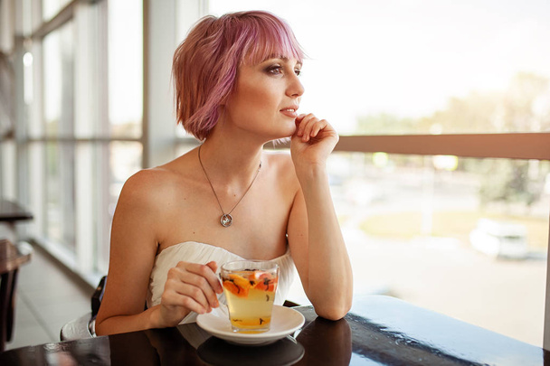 A beautiful girl with pink painted hair sits in a restaurant near the window drinking fruit tea and looking dreamily forward. Hipster girl waiting for her friend on a date - Фото, зображення