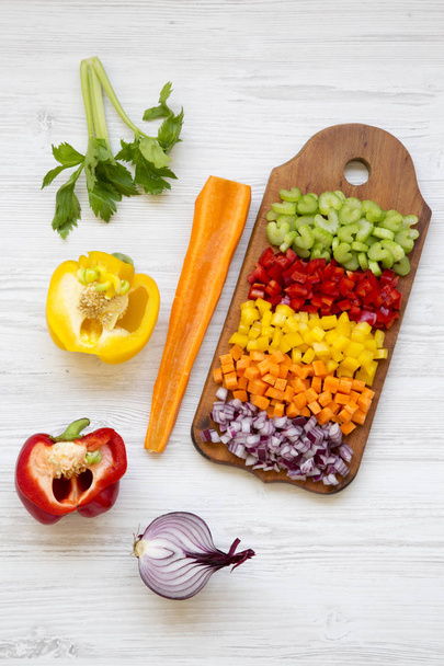 gehacktes Gemüse (Karotten, Sellerie, rote Zwiebeln, Paprika) auf Schneidebrett auf weißem Holztisch angeordnet, Blick hoch.  - Foto, Bild