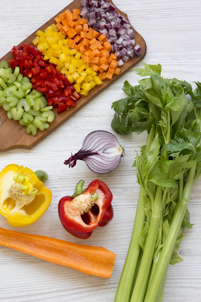 Légumes frais hachés (carotte, céleri, oignon rouge, poivrons colorés) disposés sur une planche à découper sur une table en bois blanc, vue grand angle
.  - Photo, image