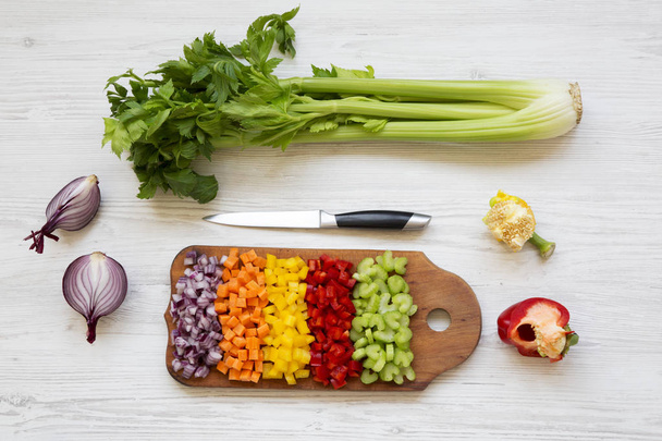 Chopped fresh vegetables (carrot, celery, onion, colored peppers) arranged on cutting board on white wooden table, overhead view. Flat lay, from above, top view. - Photo, Image