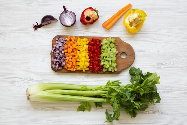Chopped fresh vegetables (carrot, celery, onion, colored peppers) arranged on cutting board on white wooden background, top view. From above, overhead. - Photo, Image
