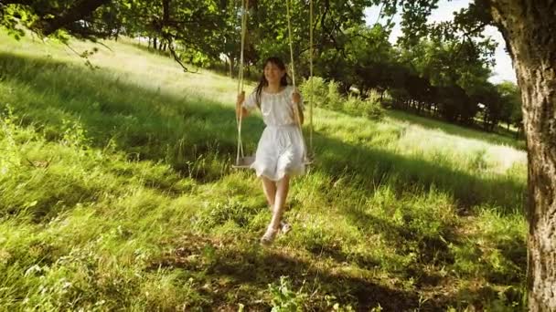 meisje met lang haar in witte jurk lacht rollen op schommel onder zomer eiken boom in een park in het najaar. Slow motion. - Video