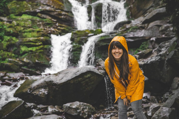 mujer de pie en impermeable amarillo y mirando a la cascada. concepto de senderismo
 - Foto, imagen
