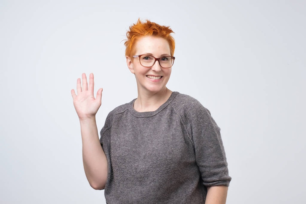 Positive mature caucasian woman with red hairstyle and glasses smiling friendly and waving hand at camera. - Photo, Image