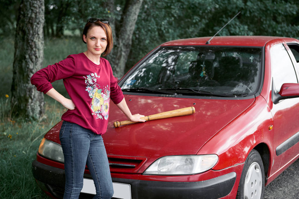 νεαρή γυναίκα οδηγός έχει άγχος και οργή, απειλεί με ένα ρόπαλο του μπέιζμπολ, έχει ένα κόκκινο αυτοκίνητο - Φωτογραφία, εικόνα