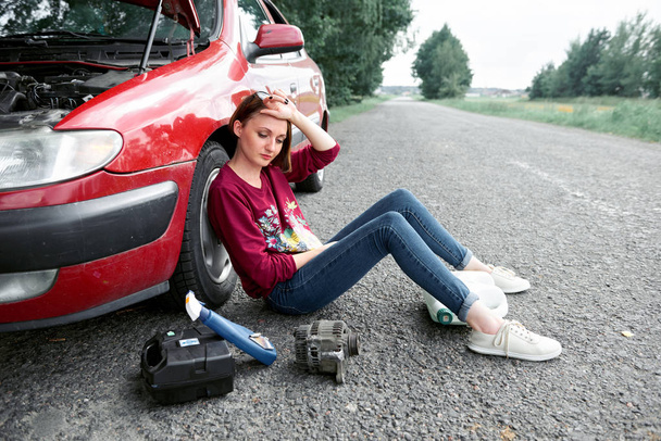 a young girl sitting near a broken car and looking for help, next to her there are bad parts, electric generator, tools and first aid kit - Photo, Image