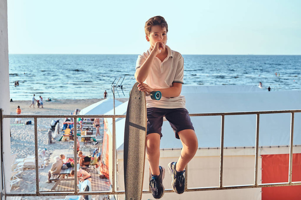 Portrait of a pensive young skater boy dressed in t-shirt and shorts sitting on the guardrail and holds a skateboard against the background of the seacoast. - Zdjęcie, obraz
