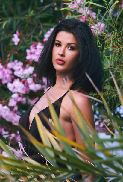 Head shot portrait of sexy young brunette woman with bob hairstyle posing outdoors. Girl pose near blooming bright beautiful pink flowers and leaves. 20-25 years old  female, looking at camera. Romantic, sensual mood, summertim - Фото, изображение