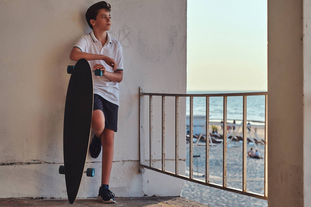 Retrato de un joven patinador vestido con camiseta y pantalones cortos apoyado en una pared y sostiene un monopatín sobre el fondo de una costa
. - Foto, imagen