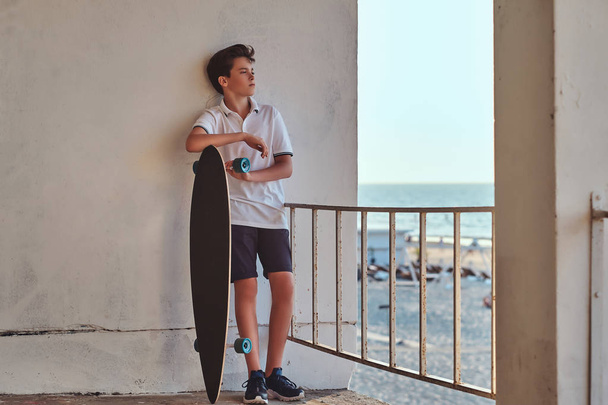 Retrato de un joven patinador vestido con camiseta y pantalones cortos apoyado en una pared y sostiene un monopatín sobre el fondo de una costa
. - Foto, imagen