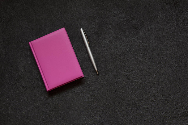 ペンとノートでオフィスのデスクトップ。黒いコンクリート背景にライラックのメモ帳。ビンテージ スタイルの抽象的なセメントの背景。トップ ビュー. - 写真・画像