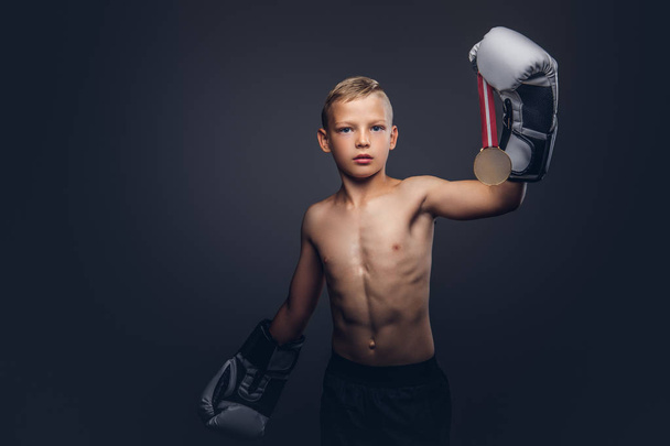 Young shirtless boy boxer in boxing gloves holds a golden medal posing in a studio. Isolated on a dark background. - Фото, изображение