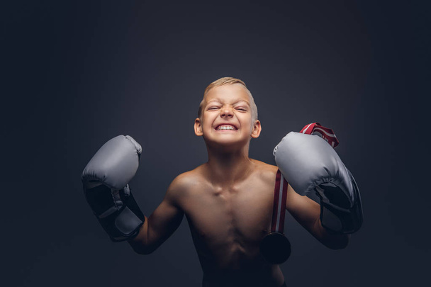 Gioioso ragazzo boxer senza maglietta in guanti da boxe tiene una medaglia d'oro in posa in uno studio. Isolato su uno sfondo scuro
. - Foto, immagini