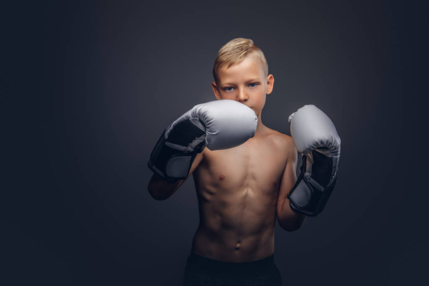 De boxer van de shirtless jongen met blonde haren dragen van bokshandschoenen training in een studio. Geïsoleerd op een donkere achtergrond. - Foto, afbeelding