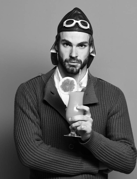 handsome bearded aviator man with long beard on serious face holding glass of alcoholic cocktail in gray knitted sweater with hat and glasses on red studio background - Foto, immagini