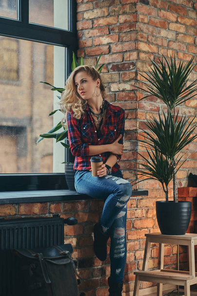 Beautiful blonde hipster girl with long curly hair dressed in a fleece shirt and jeans holds a cup of a takeaway coffee sitting on a window sill at a studio with a loft interior. - Fotoğraf, Görsel