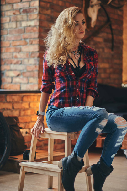 Sensual blonde hipster girl with long curly hair dressed in a fleece shirt and jeans holds a cup of morning coffee sitting on a wooden stool at a studio with loft interior. - Foto, Bild