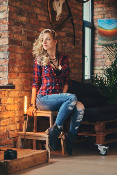 Charming blonde hipster girl with long curly hair dressed in a fleece shirt and jeans holds a cup of morning coffee sitting on a wooden stool at a studio with loft interior, looking away. - Photo, Image