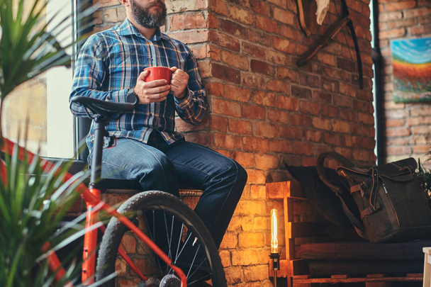 Обрезанное изображение красивого бородатого хипстера мужского пола в синей флисовой рубашке и джинсах держит чашку утреннего кофе, сидя на подоконнике в студии с интерьером лофта
. - Фото, изображение