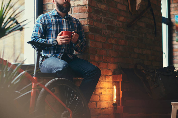 Guapo barbudo hipster macho en una camisa de lana azul y jeans sostiene una taza de café de la mañana mientras está sentado en un alféizar de la ventana en un estudio con interior loft
. - Foto, Imagen