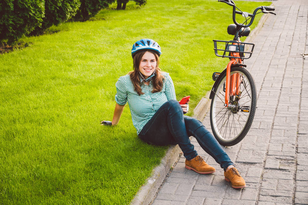 Rollendes Fahrrad für Mensch und Stadt, umweltfreundlicher Transport. Schöne junge kaukasische Arbeiterin sitzt auf dem Gras und benutzt ein rotes Mobiltelefon in der Nähe eines orangefarbenen Fahrrades mit Coryn - Foto, Bild