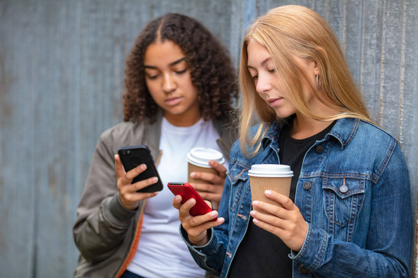 Interracial friends pretty blonde teenager and mixed race African American girl female young woman using their smart phones for social media and drinking takeout coffee outside - Fotó, kép