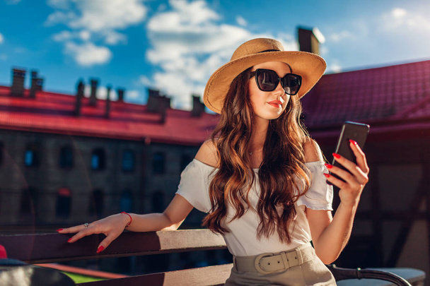 Stijlvolle jongedame hoed en zonnebril dragen en het gebruik van smartphone buitenshuis. Stijlvolle reiziger meisje ontspannen op de Bank - Foto, afbeelding