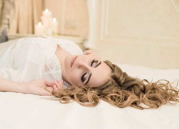Portrait of a sensual woman in white peignoir lying on the bed. Bride's morning. Beautiful lady with wedding hairstyle long curly hair and bridal makeup. - Foto, imagen