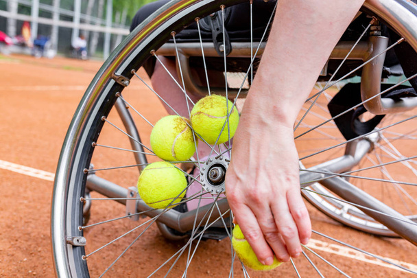 Disabled young woman on wheelchair playing tennis on tennis court. Close-up of a hand takes a tennis ball fixed in a wheel - Fotografie, Obrázek