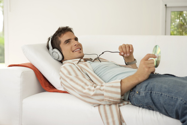 Mann legt sich auf ein weißes Sofa und hört Musik mit Kopfhörern. - Foto, Bild