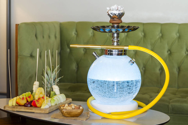 Traditional Arabic and Turkish shisha, hookah or Water pipe serving with fresh fruits, hot black tea and nuts on table, modern cafe lounge design. Led light in water. - 写真・画像