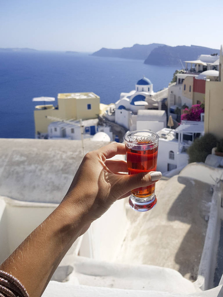 vista parcial de la mujer sosteniendo vino de rosas dulce casero en vaso pequeño con vista de la isla griega Santorini en el fondo
 - Foto, imagen