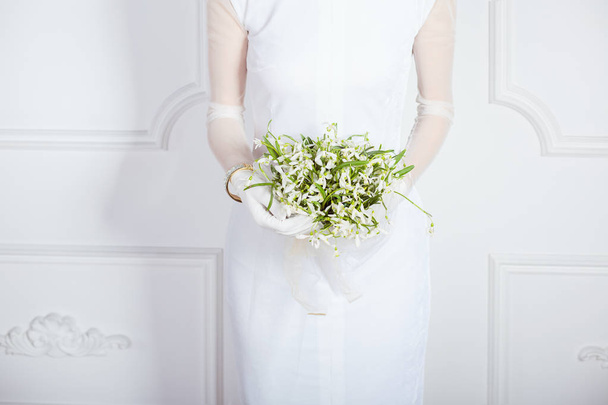 Faceless shot of bride in elegant simple dress and gloves holding small bouquet on white wall background - Fotoğraf, Görsel