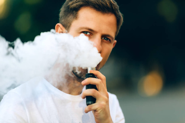 Modern young man with a beard fun Vaporizers Outdoor. Male in white t-shirt vape ecig. dark background, the evening sunset over the city. toned image. - Foto, Imagen