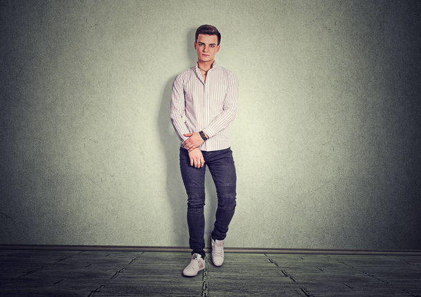 Сучасний молодий чоловік у джинсовій та білій сорочці, що стоїть з упевненістю на сірому фоні, дивлячись на камеру
 - Фото, зображення