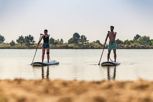 Man and woman stand up paddleboarding on lake. Young couple are doing watersport on lake. Male and female tourists during summer vacation. - Фото, изображение