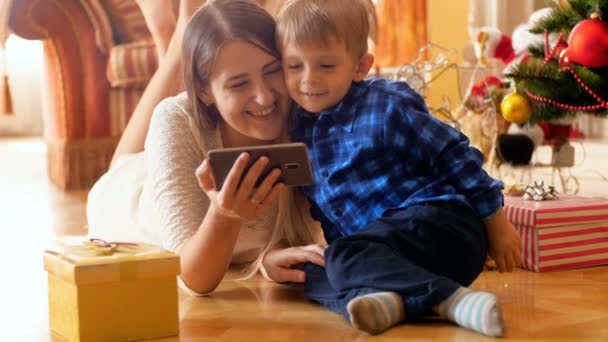 4k video of happy family lying on floor under Christmas tree and watching smart phone - Filmmaterial, Video