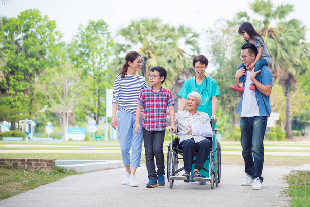 Paciente femenina mayor sentada en silla de ruedas con su familia y enfermera en el parque del hospital
 - Foto, Imagen
