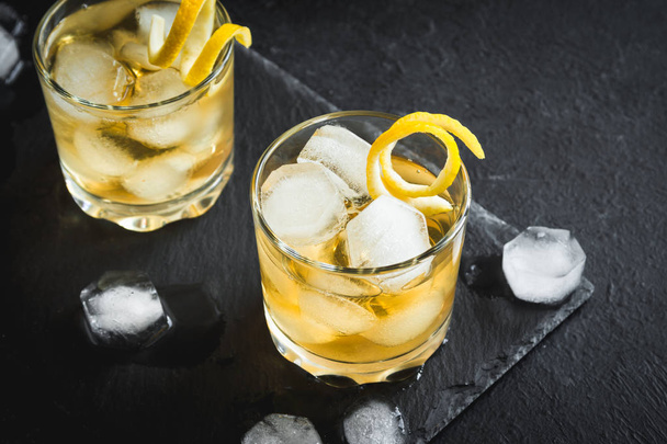 Whiskey on the rocks with lemon peel. Whiskey with ice garnished with citrus twist served on black stone background, top view, copy space. - Фото, изображение