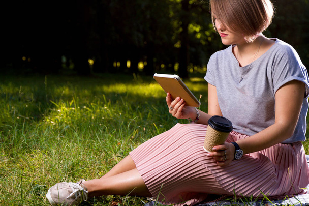 young girl sitting on plaid with coffee and reading electronic book in park - Foto, Imagen