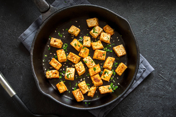 Fried tofu with sesame seeds and spices on cast iron pan, copy space. Healthy ingredient for cooking vegan vegetarian diet food. Roasted tofu over black background. - Photo, image
