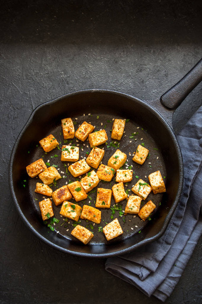 Fried tofu with sesame seeds and spices on cast iron pan, copy space. Healthy ingredient for cooking vegan vegetarian diet food. Roasted tofu over black background. - Фото, изображение
