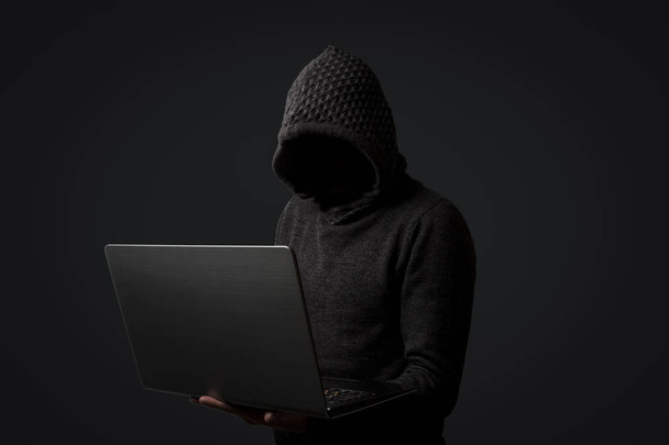 Egy kapucnis pulcsit, kapucnival arctalan ember rendelkezik a laptop a kezében a sötét háttér előtt. Szaggató köhögés és lopás felhasználói adatok fogalmának. - Fotó, kép