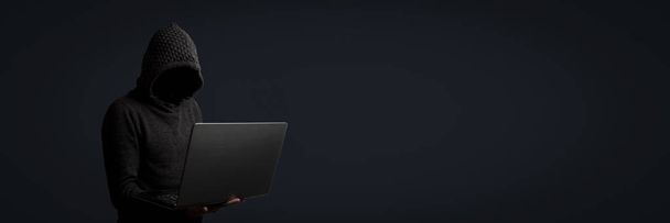 Un hombre sin rostro con capucha y capucha sostiene un portátil en sus manos sobre un fondo oscuro. Concepto de hackeo, robo de datos de usuarios. Cerradura hackeada, tarjeta de crédito, nube, correo electrónico, contraseñas, archivos personales. Banner
. - Foto, imagen