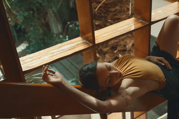 Екзотична жінка відпочиває, лежачи на дерев'яних сходах поруч з вікном з пальмами, тропічним садом на задньому плані. Мрійлива леді
 - Фото, зображення