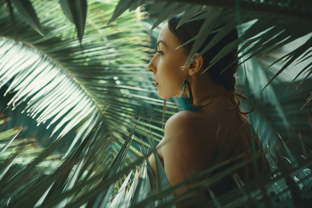 Portrait exotic woman with naked shoulders and big earrings with soft feathers touching with hand her skin while relaxing among tropical plants. Copy space on right. Light play on palm leafes. - Photo, Image